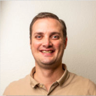 A man in a tan shirt smiling in front of a white wall.